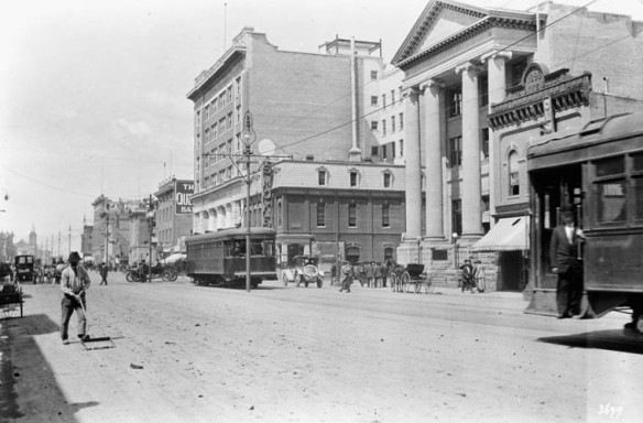 A black-and-white photograph showing a wide avenue, roughly paved, where streetcars, horse-drawn carriages and automobiles share the road. It is a streetscape bustling with activity.