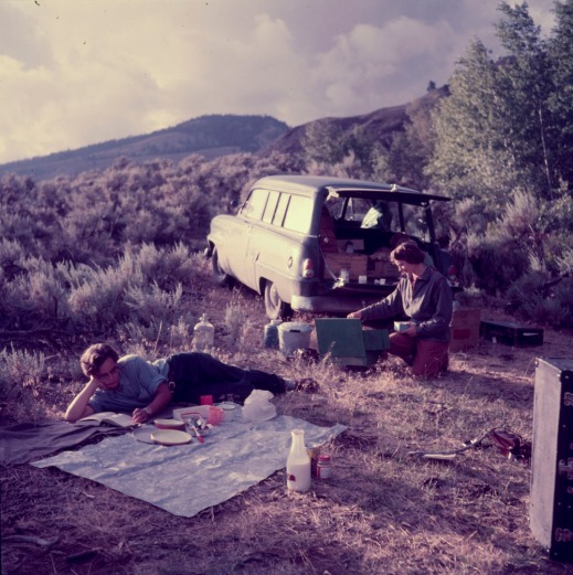 A colour photograph of two women in a grassy area with mountains in the distance—one is reading reclined on a picnic blanket and the other is kneeling at a camp stove located behind a station wagon.