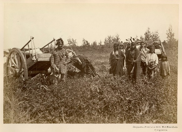 Black and white photograph of a man, on the left, wearing European clothing and standing in front of a Red River cart, and a group of First Nations men, women and children wearing First Nations-style clothing and standing in front of another Red River cart, on the right.