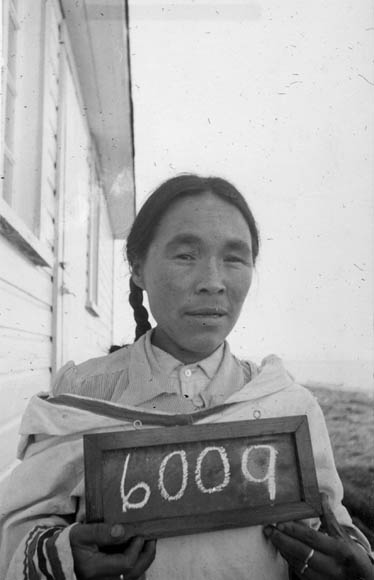 A black-and-white photograph of an Inuit woman holding a small chalkboard with the number 6009. 