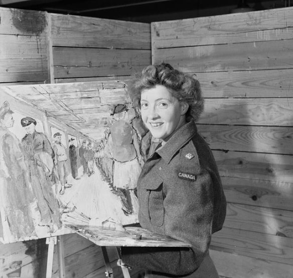A black-and-white photograph showing Molly Lamb Bobak posing in front of an easel with brushes and palette in hand. Bobak wears an army battledress jacket and smiles at the camera. The partially completed painting behind her depicts male and female members of the Canadian Army standing inside a room.