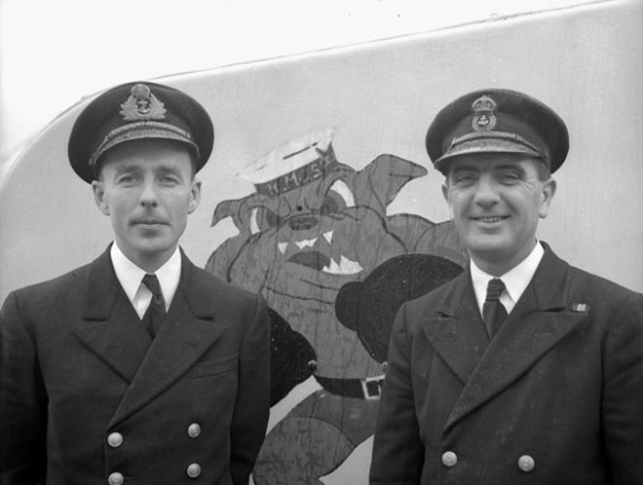 Black-and-white photograph, showing two men in naval uniform posing in front of the nose turret of their corvette. Between the two men, an image painted on the turret shows a bulldog standing on his hind legs, wearing a sailor hat and boxing gloves. 
