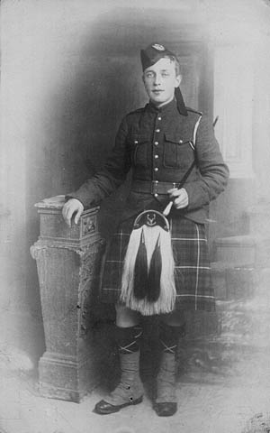 A black-and-white photograph of a young man wearing a kilt and sporran, holding a baton in his left hand and leaning on a sculptural shelf.