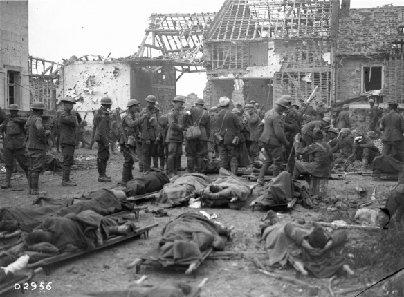 A black-and-white photograph showing a crowded war scene: wounded soldiers are on stretchers while soldiers mill around, with destroyed buildings in the background. 