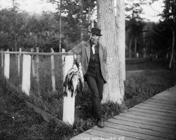 A black-and-white photograph of a man wearing a hat, coat and tie leaning against a fence post and holding a string of fish.
