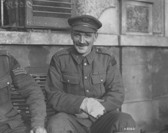 A black-and-white photograph of a seated soldier with a bandaged hand smiling at the photographer. 