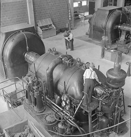A black-and-white photograph of a man on a small platform examining the pressure gauge of a turbine steam generator.