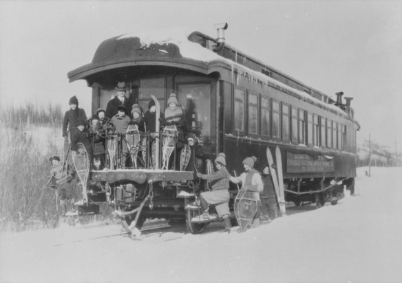 A black-and-white photograph of thirteen children and their teacher posing with their snowshoes at the entry deck of a Canadian National Railway school car.