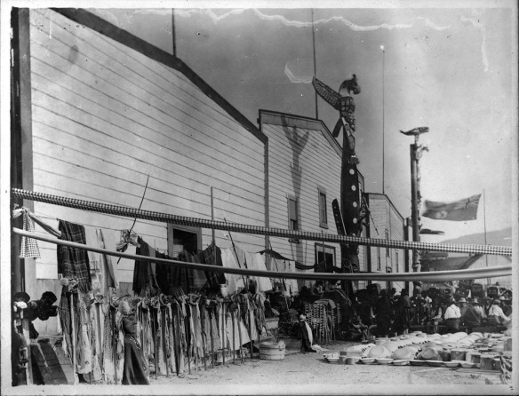 A black-and-white photograph of a streetscape with potlatch participants and items to be given away.