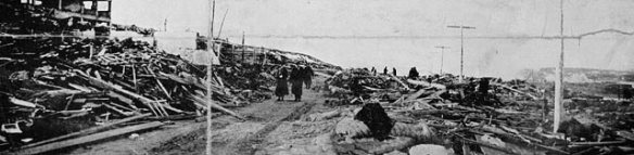 A black-and-white photograph of several people walking down a street with destroyed buildings on both sides.