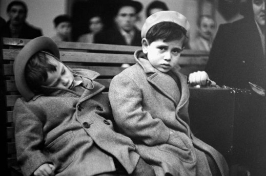 A black-and-white photograph of two small boys wearing wool coats and hats sitting on a wooden bench. One is slumped over sleeping; the other is staring the camera and holding a suitcase. A blurred crowd of people can be seen in the background.