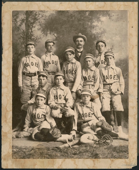 A black-and-white photograph of 10 children wearing baseball uniforms. The jerseys read "Pages" across the front. The boys are sitting and standing with bats, gloves and other baseball equipment. Behind the boys stands an adult man, wearing a suit and hat. The background is a studio backdrop showing trees.