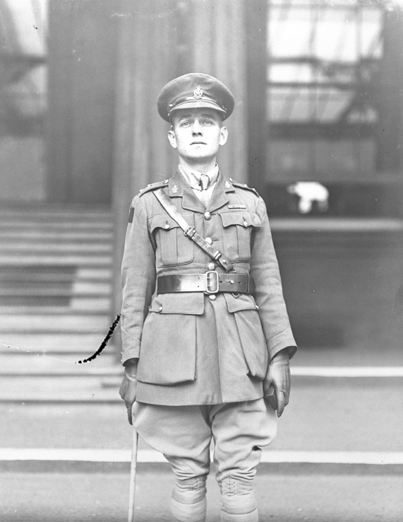 A black-and-white photograph of a soldier in an officer’s uniform with gloves and a cane standing in front of stairs and a window.