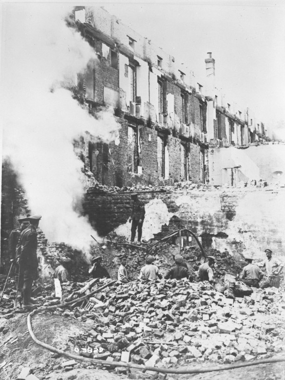 A black-and-white photograph of uniformed soldiers working amidst the rubble of a large, heavily damaged brick building that has been bombed.