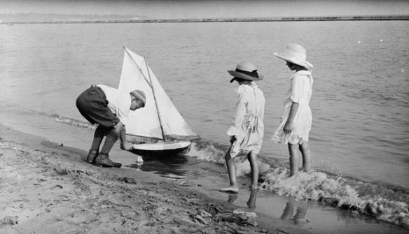 A black-and-white photograph of two girls watching a boy launch a model yacht into the water. 