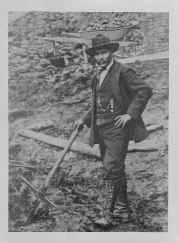 A black-and-white photograph of a man holding some prospecting equipment with one hand and the other hand on his hip looking directly at the viewer. Behind him is a loaded wheelbarrow.