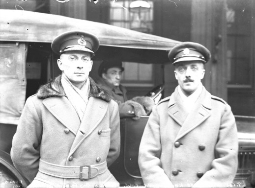 A black-and-white photograph of two officers standing in front of a car in which a driver is seated. They both wear officers’ caps and greatcoats.
