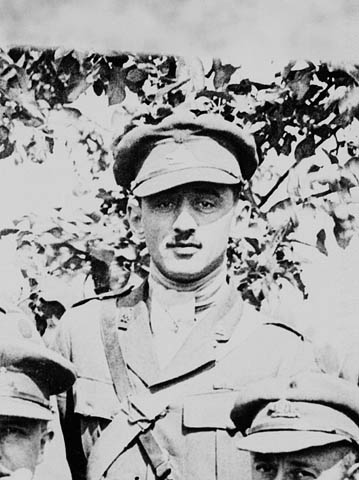 A black-and-white photograph of a soldier in uniform looking straight at the camera. He is standing behind two other men in uniform whose faces are partially visible in the foreground. There is a tree in the background. 