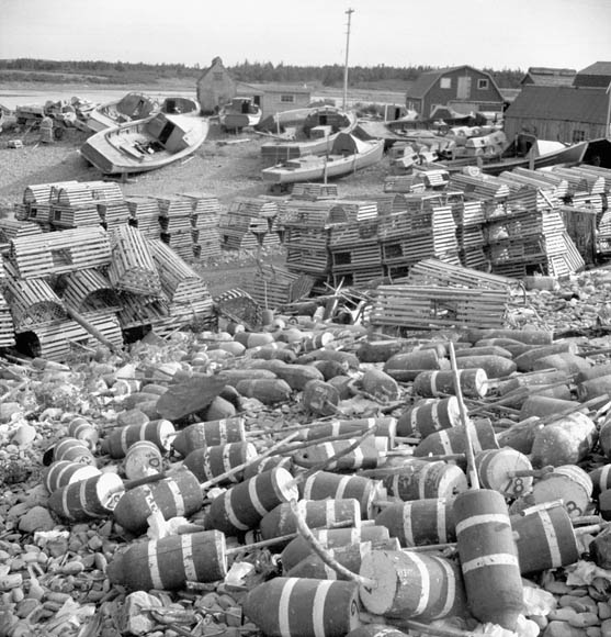 A black-and-white photograph of a coastal village, with lobster boats in the background, lobster pots in the middle distance, and floating markers in the foreground. 