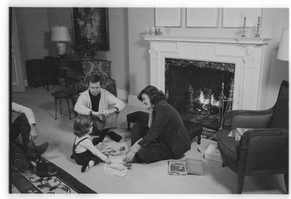 Black-and-white photograph depicting Prime Minister Joe Clark with his wife and daughter, sitting on the floor in the living room, in front of a fireplace.