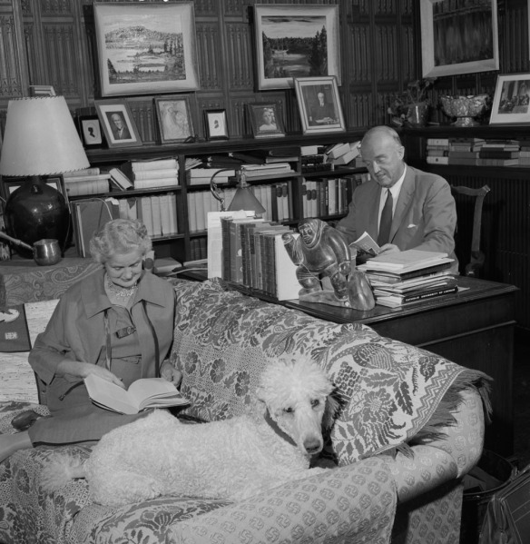 A black-and-white photograph of a woman sitting on a couch reading a book beside her poodle. A man sitting at a desk next to the couch reads a magazine. 