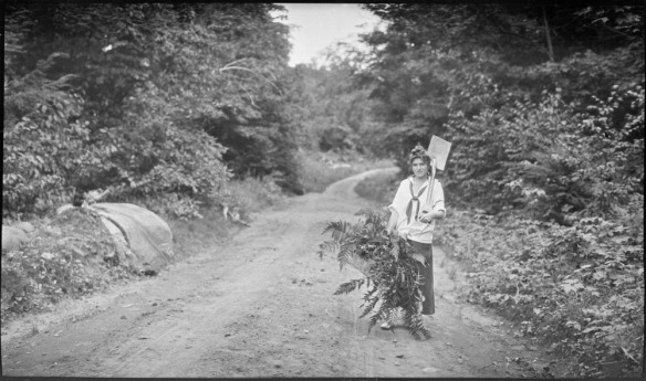 Black-and-white photograph of a woman standing on a dirt road holding a basket full of fern leaves and a shovel on her left shoulder.