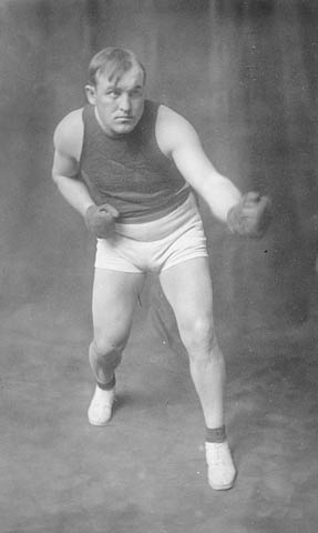 Black-and-white photo of a man wearing boxing gloves, shorts and shoes.