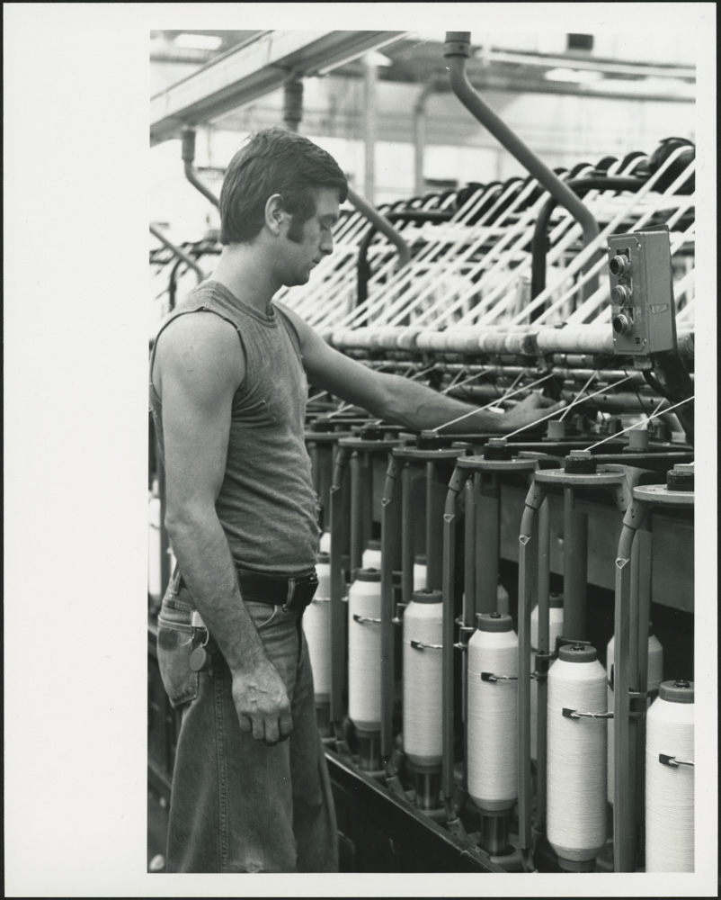 A black-and-white photograph of a man wearing jeans and a sleeveless T-shirt monitoring a spooling machine.