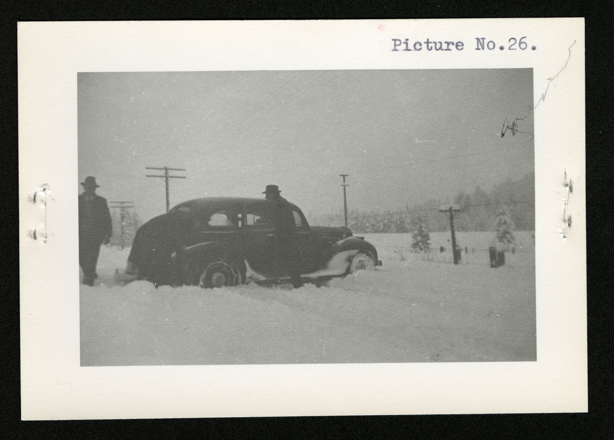 Three men and a car in a snowstorm: (from left to right) one man standing at the rear of the car, a second man bent over the back right tire, and a third man going towards the car to assist.