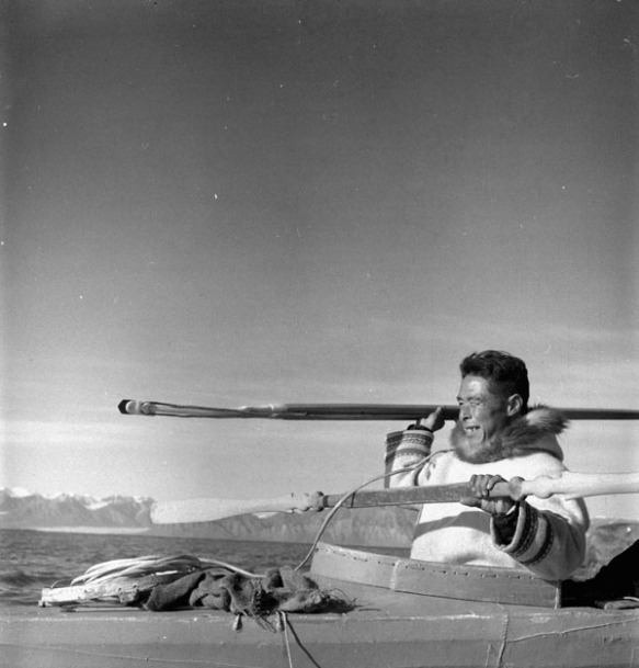 A black-and-white photograph showing a man in a qajaq about to throw a harpoon. There are snow-covered mountains in the distance.