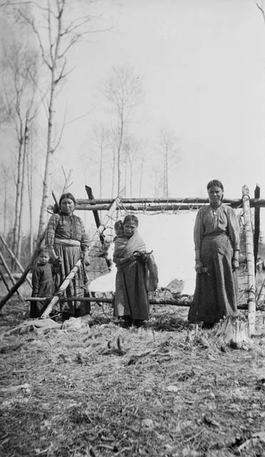 Black-and-white photograph of two women and a girl standing outdoors in front of a stretched moosehide laced onto a wooden frame. The girl is carrying an infant, and the woman on the left is holding the hand of a small child. They are on grassy vegetation, and there is a row of tall deciduous trees without leaves in the background.