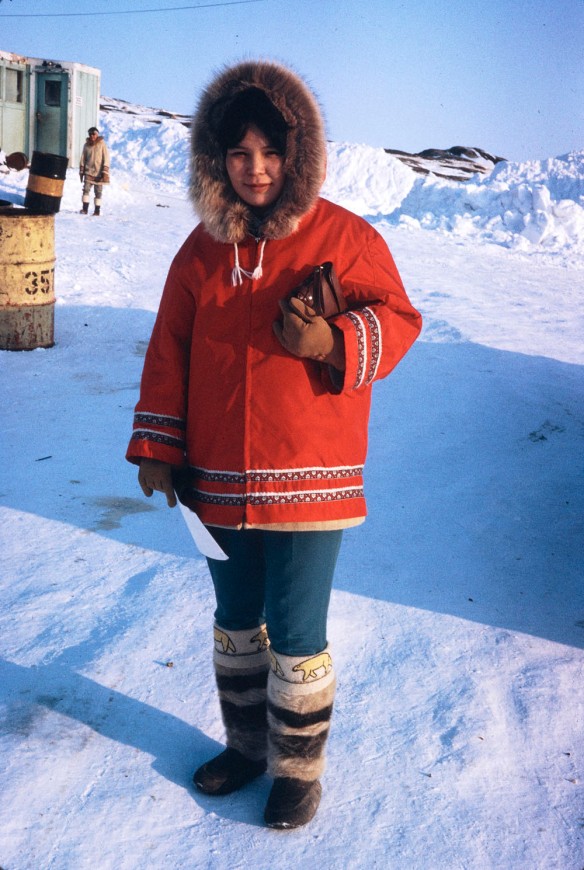 Photograph of a woman standing outside on snow-covered ground. She is wearing a red parka with a fur-trimmed hood covering her head, and knee-high boots with fur and embroidered polar bears. She is holding a brown leather purse with her left arm.