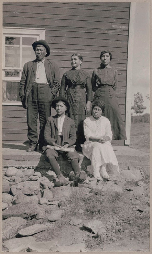 Husband and wife standing outside a building with their daughter standing next to her mother and their son and second daughter sitting in front of them.