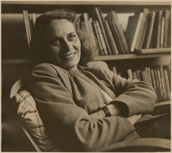 Sepia photograph of a Caucasian woman with dark hair, smiling and sitting in front of a bookshelf with her arms crossed.