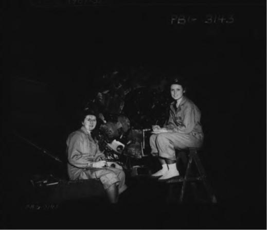 Two women wearing coveralls sit on either side of a propellor.
