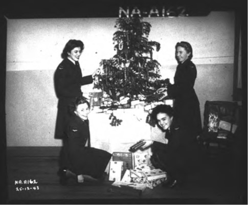 Four uniformed women stand and kneel around a Christmas tree positioned atop a table. Wrapped gifts appear on the floor and underneath the tree.
