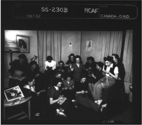 A group of fifteen women, mostly in civilian dress, engages in various leisure pursuits in what looks like a living room or lounge-like setting. Many appear to be engaged in needlecraft.